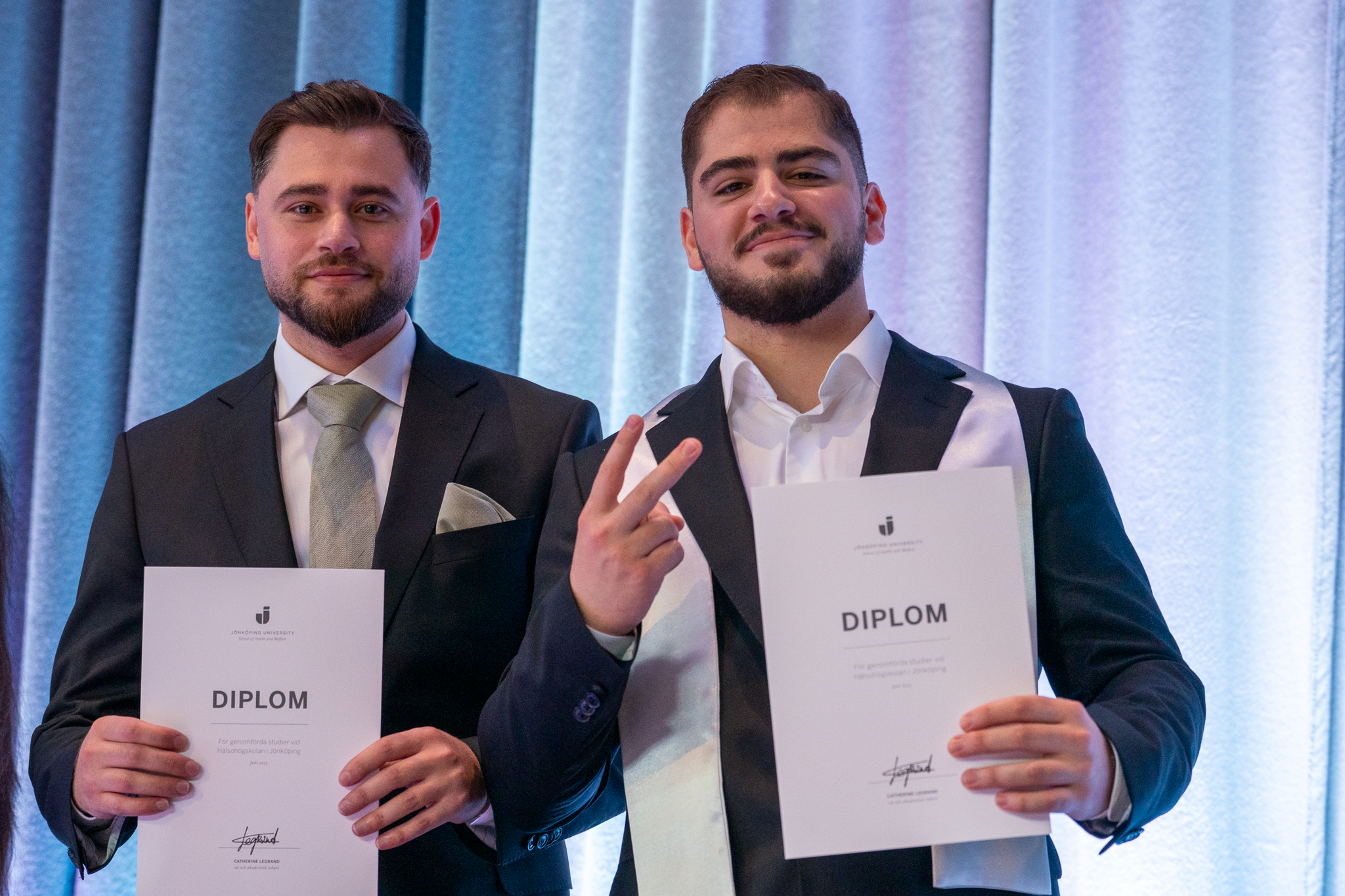 Two guys holding their diplomas and looking at the camera.