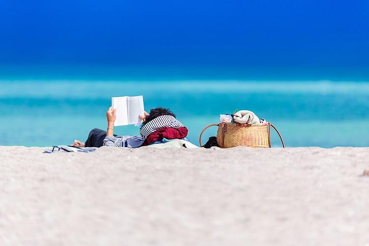 En person ligger på en sandstrand och läser en bok. Bredvid står en väska och framför syns havet.