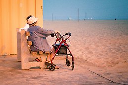 Äldre dam med rollator sitter på en bänk i solig miljö