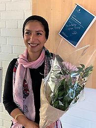 Sumaya Hashim stands smiling out at the camera, she is holding a bunch of flowers and standing in front of a wooden board where her thesis is hanging up by a nail. 