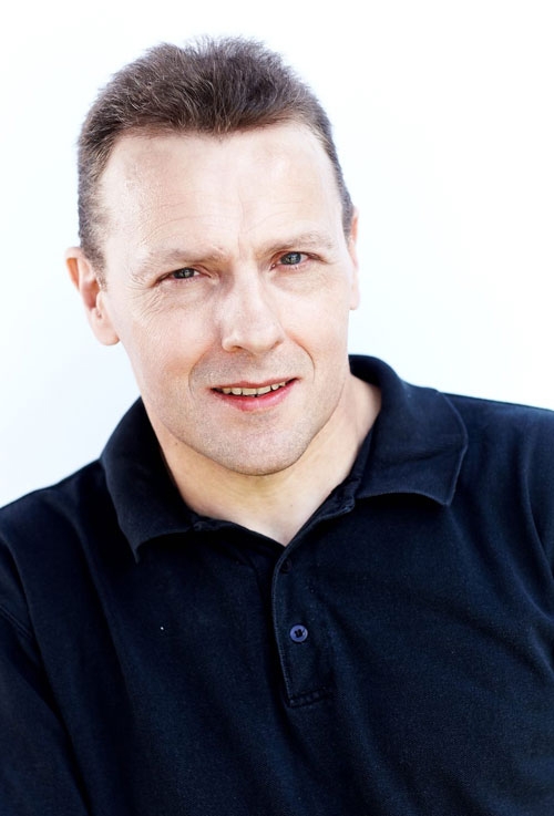 Man wearing a black collared shirt, facing forward and smiling slightly, with short hair and a light background.