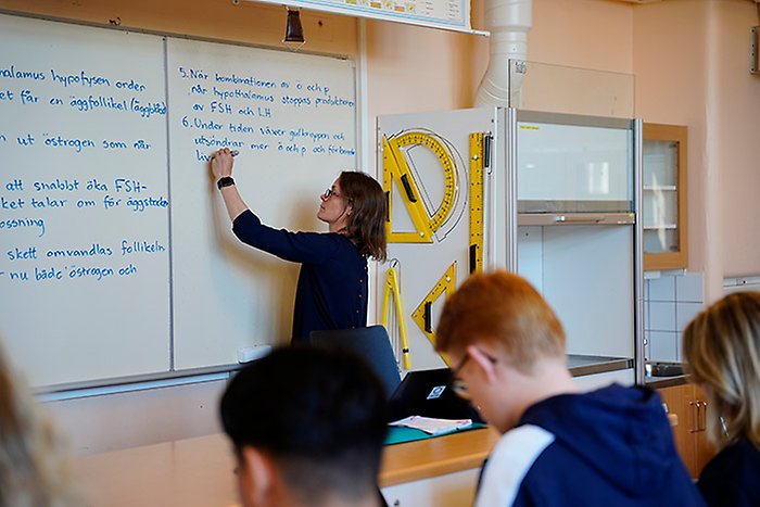 Lärare skriver på tavlan, några barn i förgrunden.